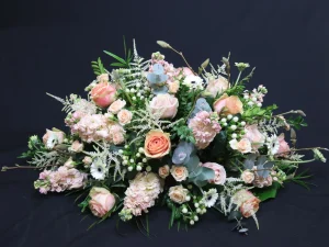 Peach Flowers, een rouw bloemstuk met verschillende bloemen in pastelkleuren zoals zachtroze en lichtblauw, afgewerkt met groen. Dit is een langwerpig en hoog rouwstuk, gemaakt door Bloeibloemen.nl