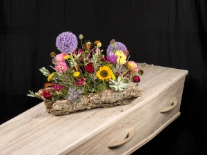 Robust color, een rouw bloemstuk op een kist met bloemen in de kleuren paars, geel, rood en roze op een boomschors, gemaakt door Bloeibloemen.nl