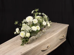 Robust white, een rouw bloemstuk op een kist met bloemen in witte kleuren op een boomschors, gemaakt door Bloeibloemen.nl