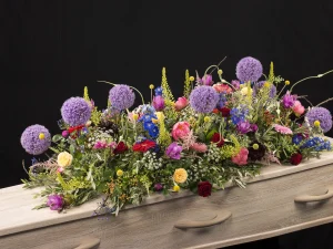 Sfeer kleuren, een rouw bloemstuk op een kist met bloemen in de kleuren paars, roze, geel, rood, blauw en groen, gemaakt door Bloeibloemen.nl