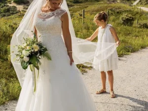 Een bruid en een bruidsmeisje die de sluier vasthoudt, ze staan in de duinen. De bruid heeft een bruidsboeket in haar handen.