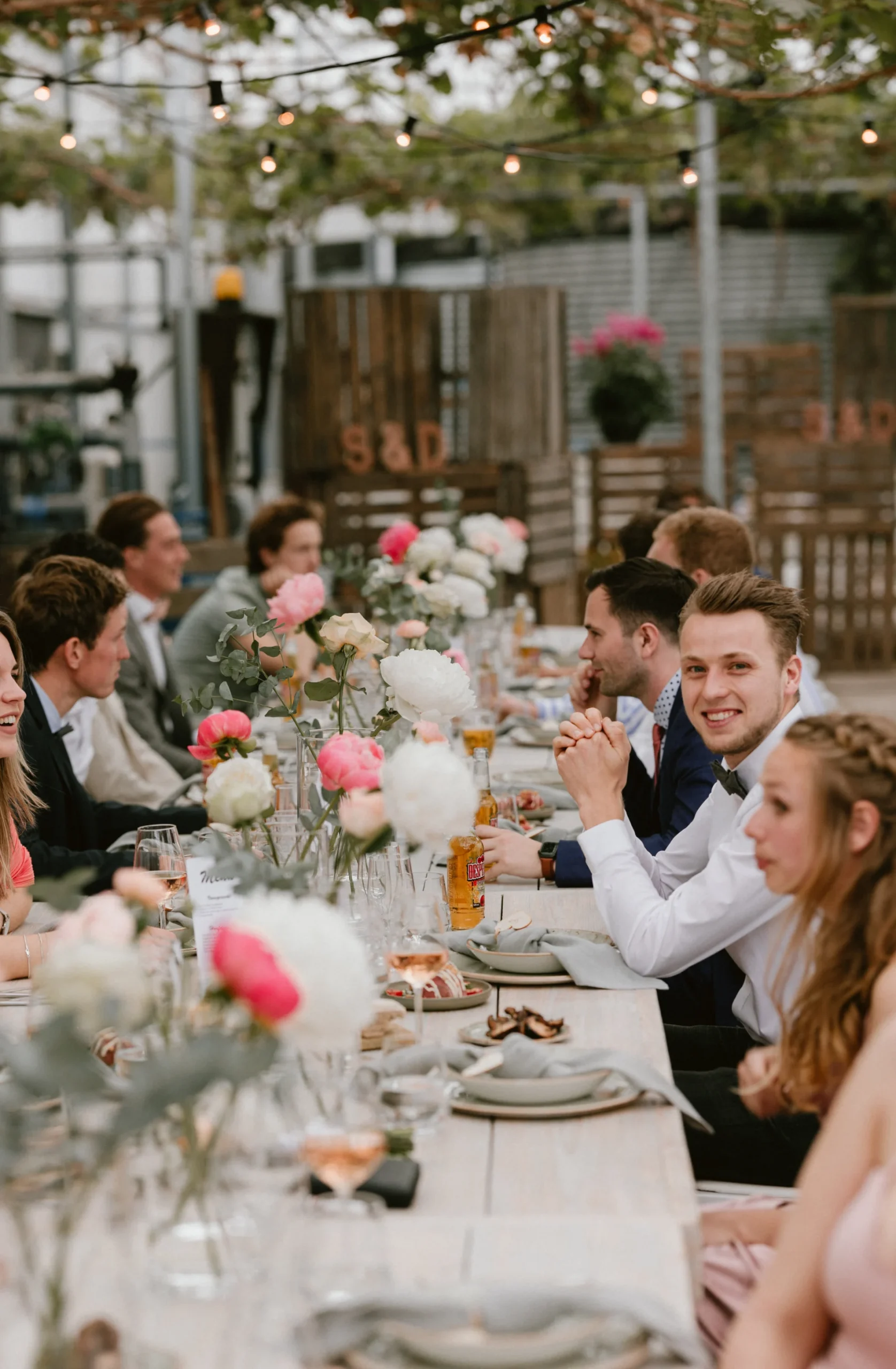 Gedekte tafel bij een bruiloft met mensen aan tafel. Op de tafel staan tafelvaasjes met bloemen met bloemen in de kleuren wit en roze.