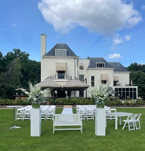 Een foto van een buiten trouwlocatie met bloemen, stoelen en een groot landhuis op de achtergrond