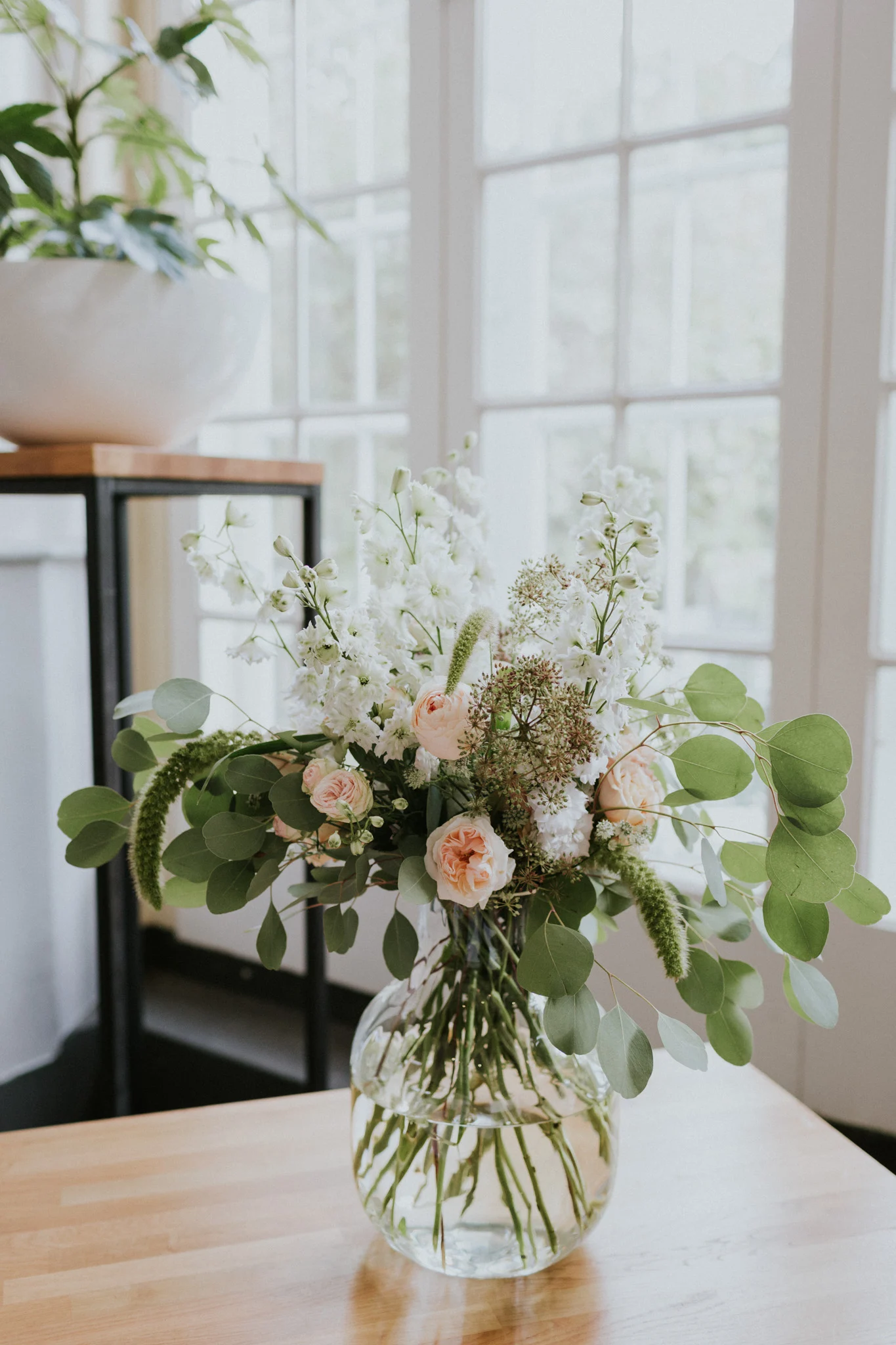 Een boeket met bloemen in een transparante vaast. De bloemen hebben de kleuren wit en zachtroze. Mooi om thuis te laten bezorgen.
