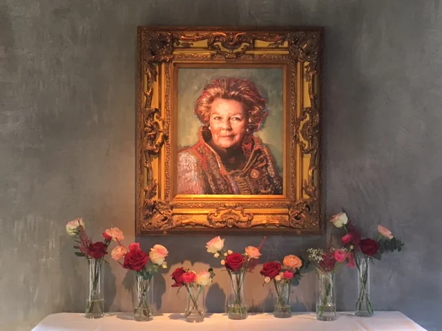 7 vaasjes met bloemen in de kleuren roze en rood onder een getekend schilderij van Beatrix.