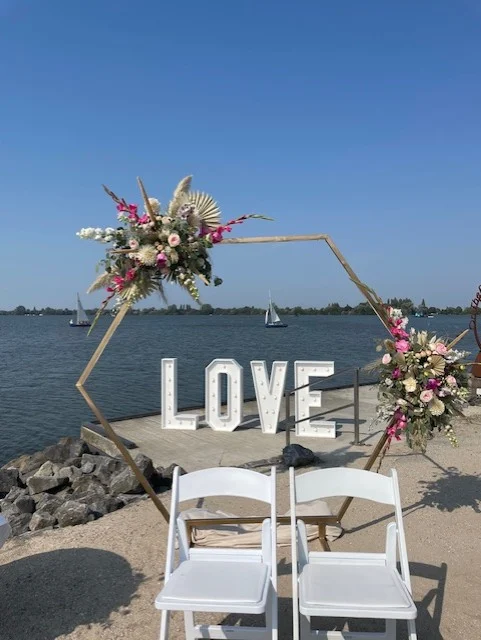 Een zuil met twee grote bloemstukken erop met daartussen grote letters met Love. Voor de zuil staan twee stoeltjes waar het bruidspaar op komt te zitten tijdens de ceremonie.
